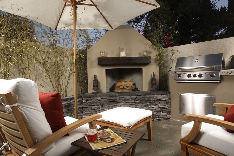 Outdoor patio with fireplace, grill, wooden furniture with white cushions, umbrella, and Asian-inspired statues.