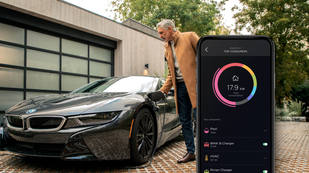 A man in a beige coat connects a charger to a car parked in front of a modern home, with a smartphone displaying an energy consumption chart on its screen.