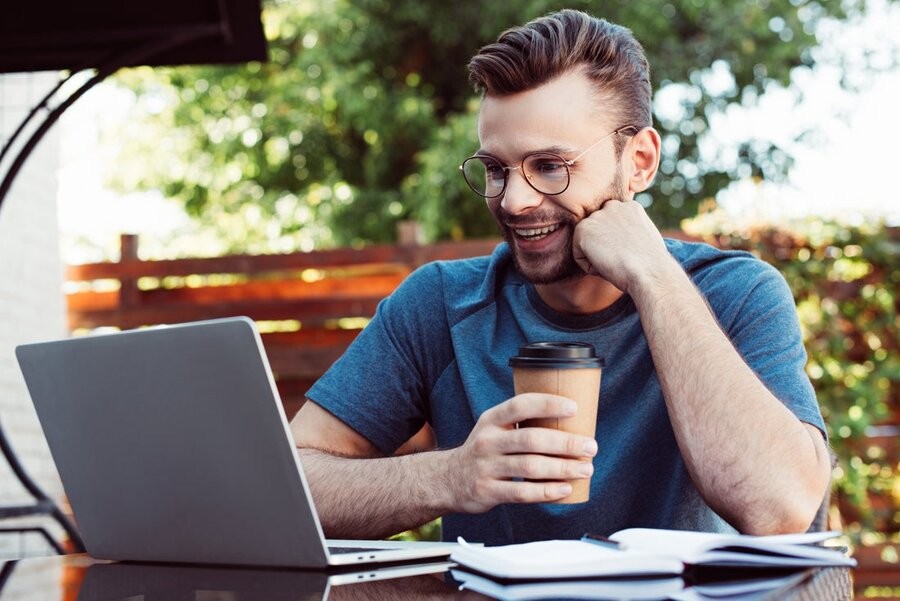 A man outside in a patio area on his laptop at a table while holding a coffee.