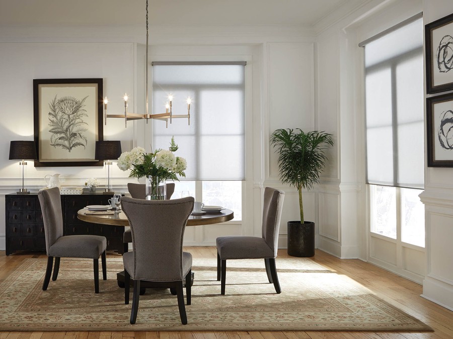 A small dining area with a table and four chairs with windows in the background featuring motorized shades. 